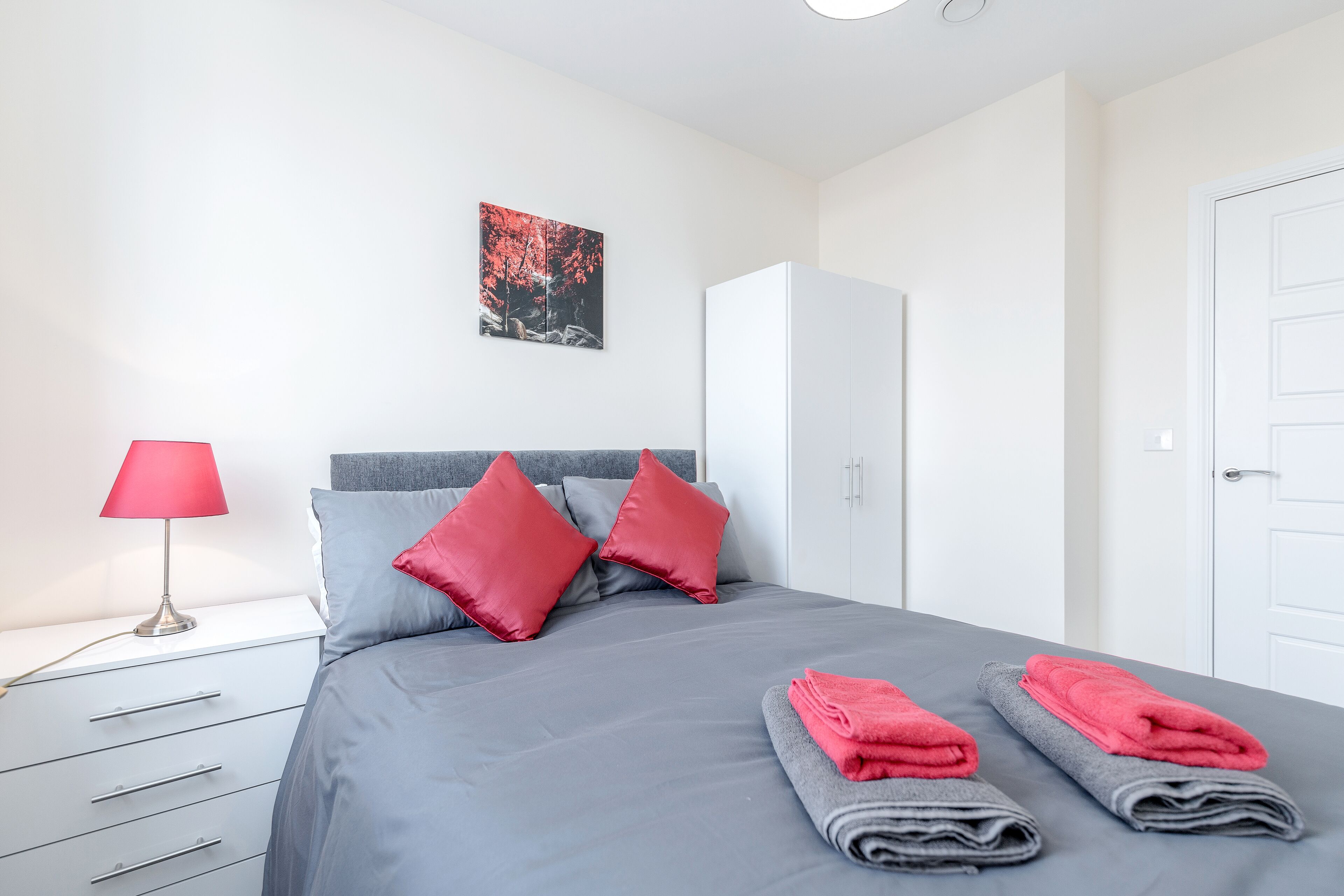 A neatly made bed with grey and red bedding, a white wardrobe, and a red lamp on a bedside table.