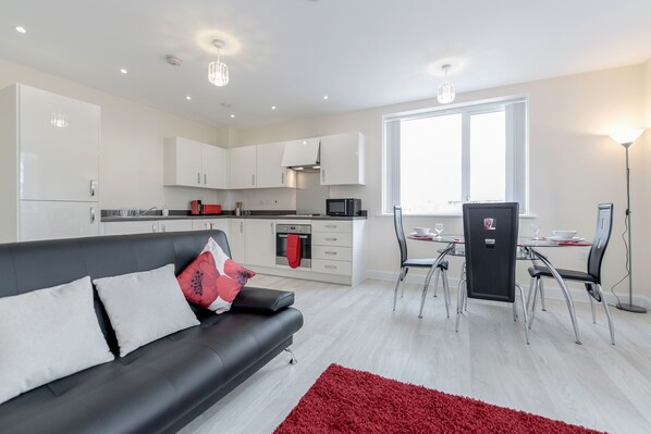 A modern kitchen with white cabinets, a dining area with a glass table, and a black sofa with red cushions.