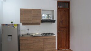A compact kitchen with wooden cabinets, a built-in gas stove, a refrigerator, and a window with blinds. - Laureles Apartments! (Medellín)