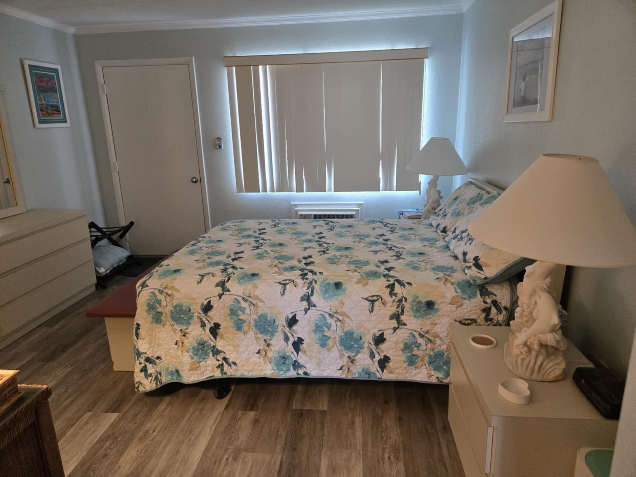 A bedroom with a floral bedspread, a bedside lamp, a dresser, a chair, and a window with blinds.