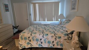 Un dormitorio con colcha de flores, lámpara de noche, cómoda, silla y ventana con persianas.