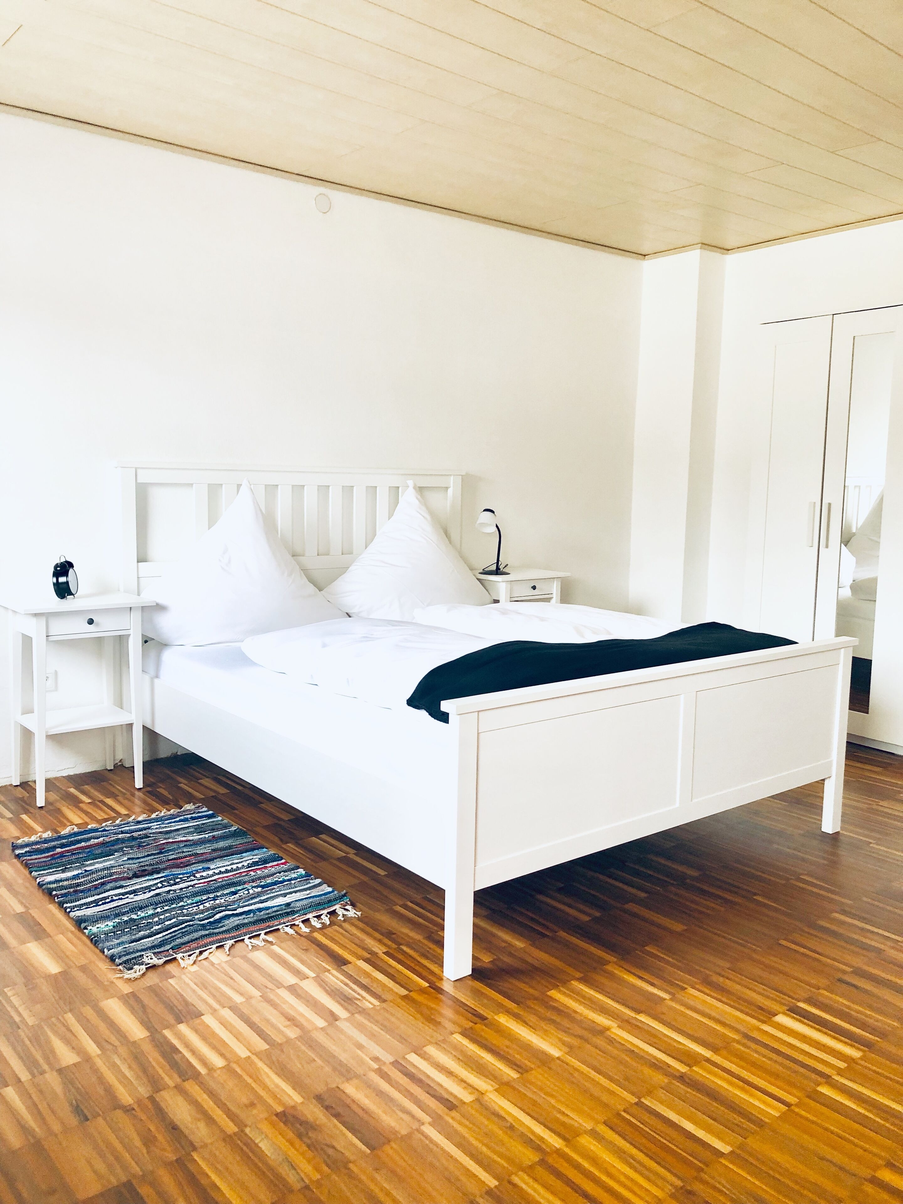 Un dormitorio con una cama blanca, una mesita de noche, un armario grande y suelo de madera.