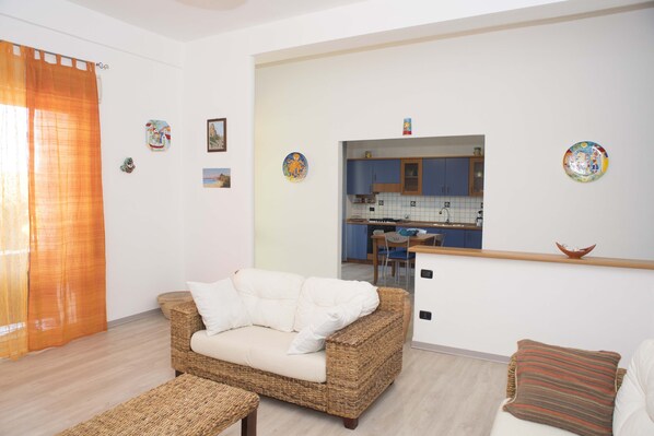 A living room with a wicker sofa, a small table, and a kitchen in the background. - Villa Eden Luxore (SCIACCA)