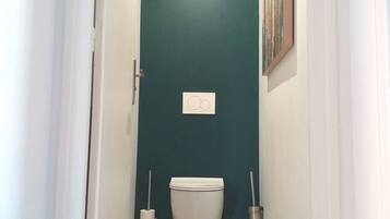 Un couloir étroit, avec des toilettes au fond, une porte sur le côté gauche et un tableau accroché au mur de droite.
