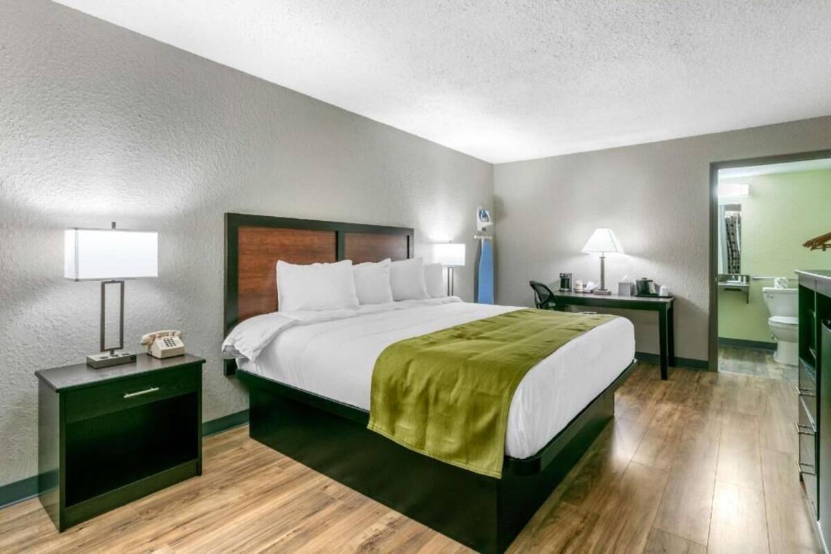 A hotel room with a bed, bedside tables, a nightstand, a lamp, and a bathroom visible through an open door.