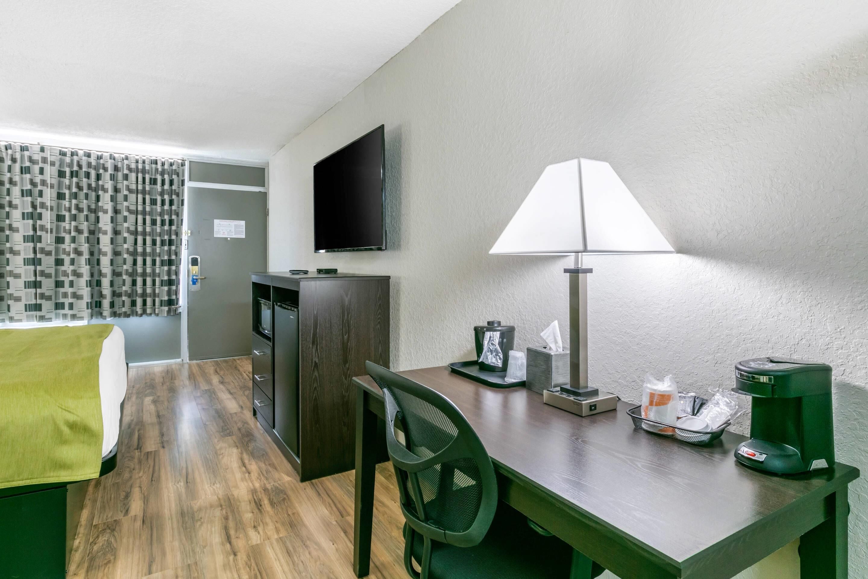 Ein Hotelzimmer mit Bett, Schreibtisch, Stuhl, Fernseher, Lampe, Kaffeemaschine und einem kleinen Tisch mit diversen Gegenständen.
