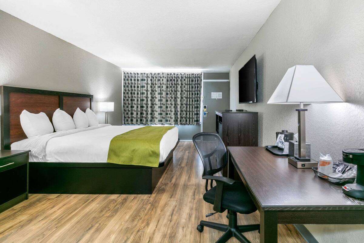 A hotel room with a bed, desk, chair, lamp, TV, and a curtain.
