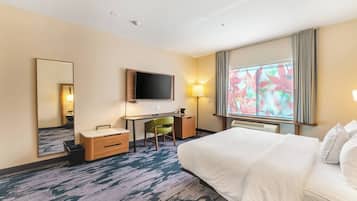Habitación de hotel con una cama grande, un escritorio con silla, un televisor de pantalla plana, un espejo y una ventana con vistas a hojas otoñales.