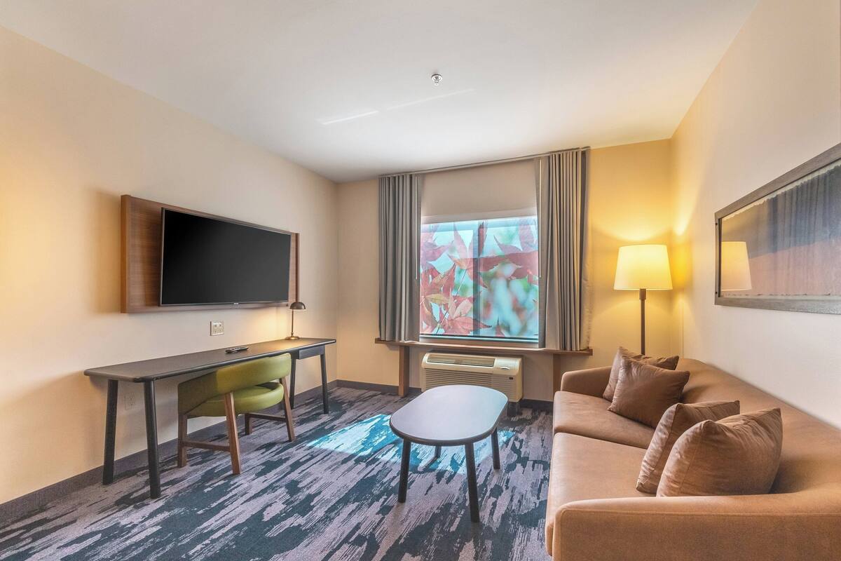 Une chambre d’hôtel équipée d’une télévision à écran plat, d’un bureau avec une chaise, d’un canapé, d’une petite table, d’une lampe et d’une fenêtre avec des rideaux.