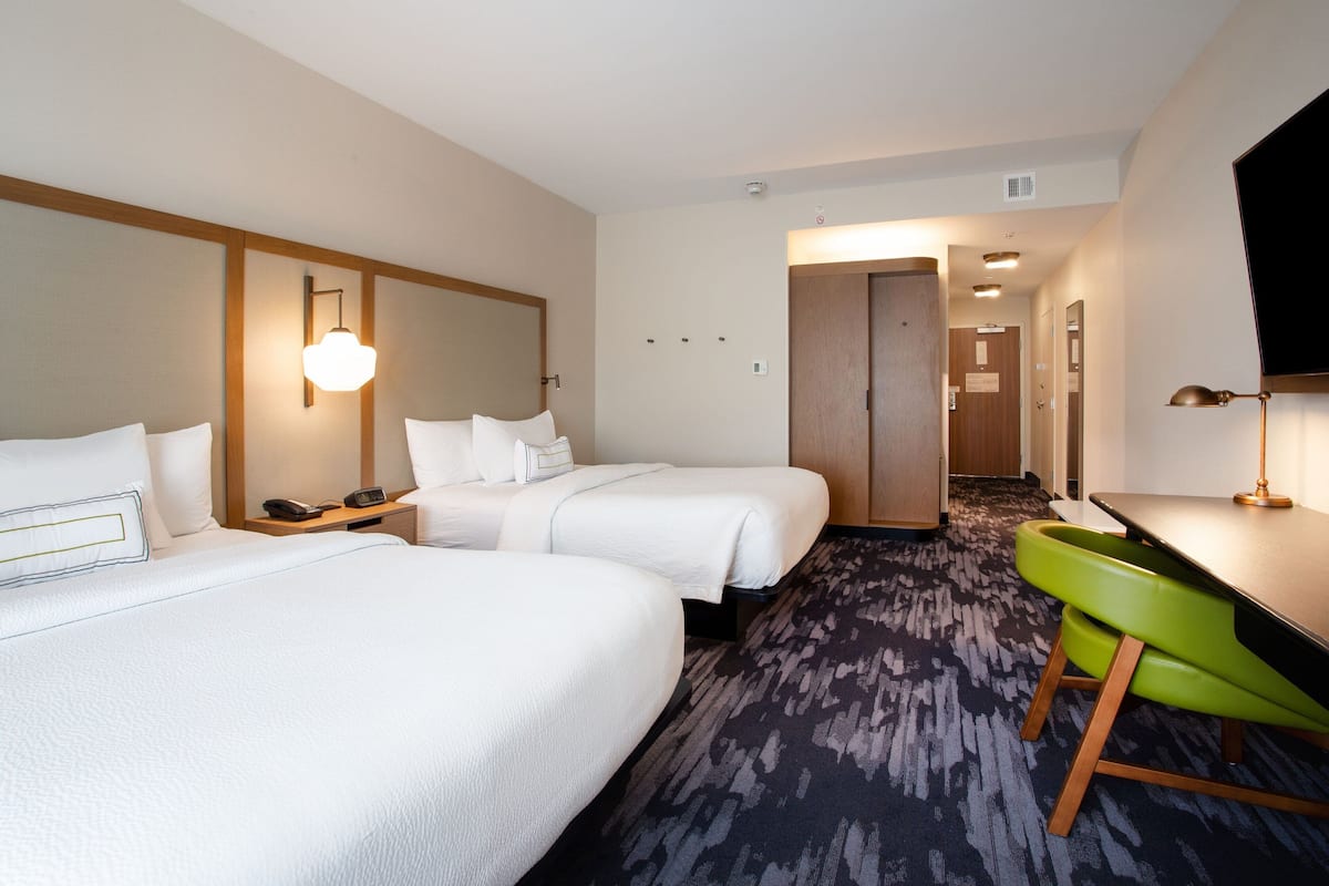 Quarto de hotel com duas camas, uma escrivaninha, uma cadeira verde e uma televisão.