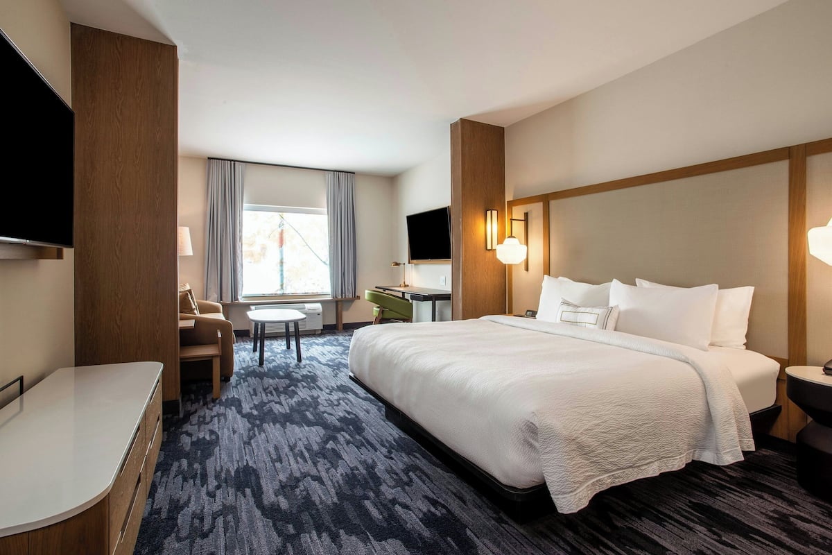 Quarto de hotel com uma cama grande, uma escrivaninha, uma cadeira, uma televisão e uma janela com cortinas.