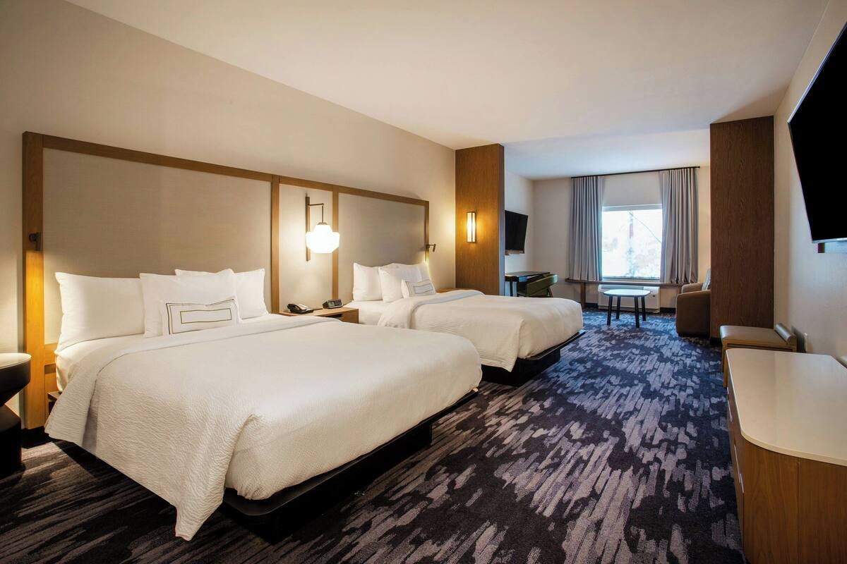 Quarto de hotel com duas camas, uma escrivaninha, uma cadeira, televisão e janela com cortinas.