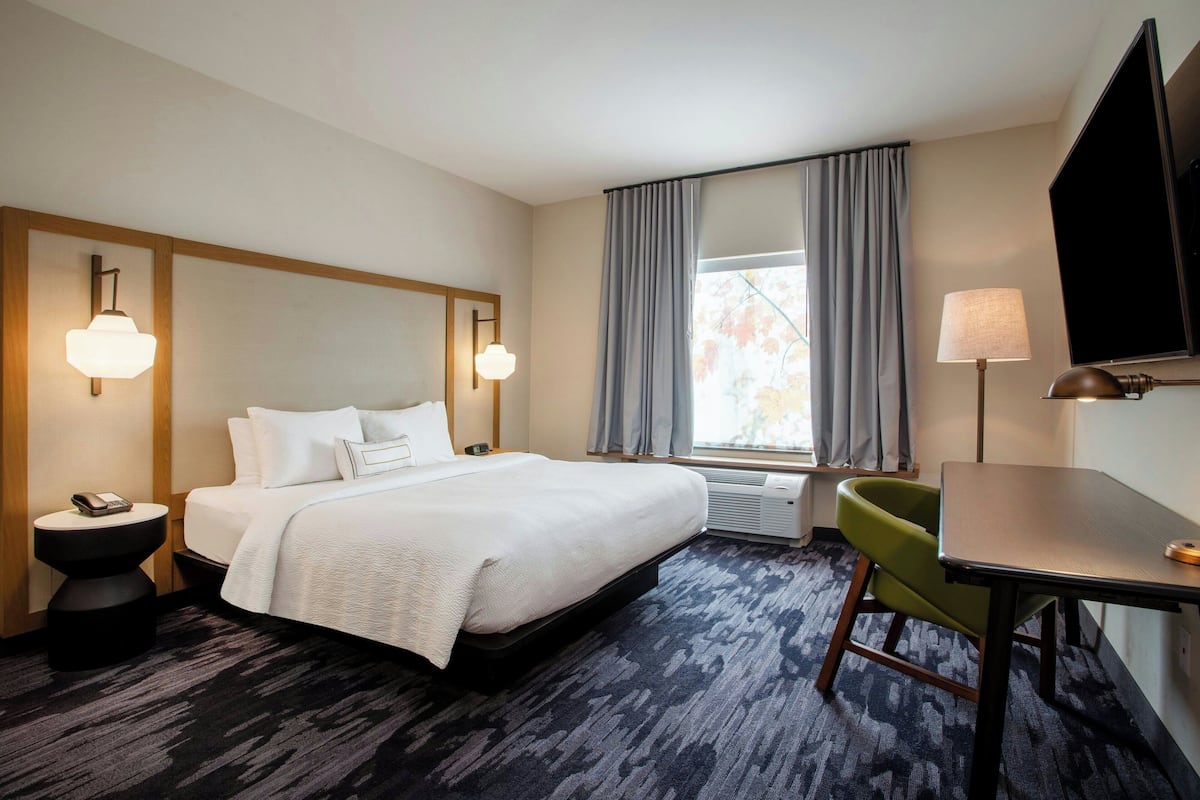 Quarto de hotel com uma cama grande, uma escrivaninha, uma cadeira, uma televisão e uma janela com cortinas.