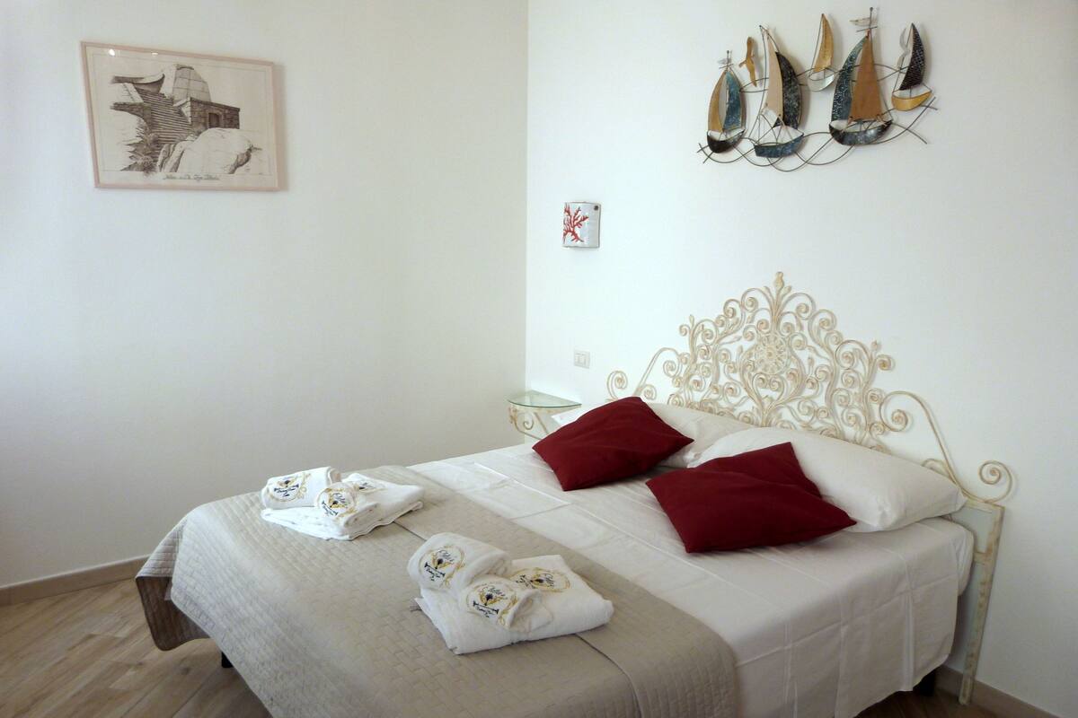 A neatly made bed with white linens and red pillows, a framed artwork on the wall, and a decorative wall hanging of sailboats.