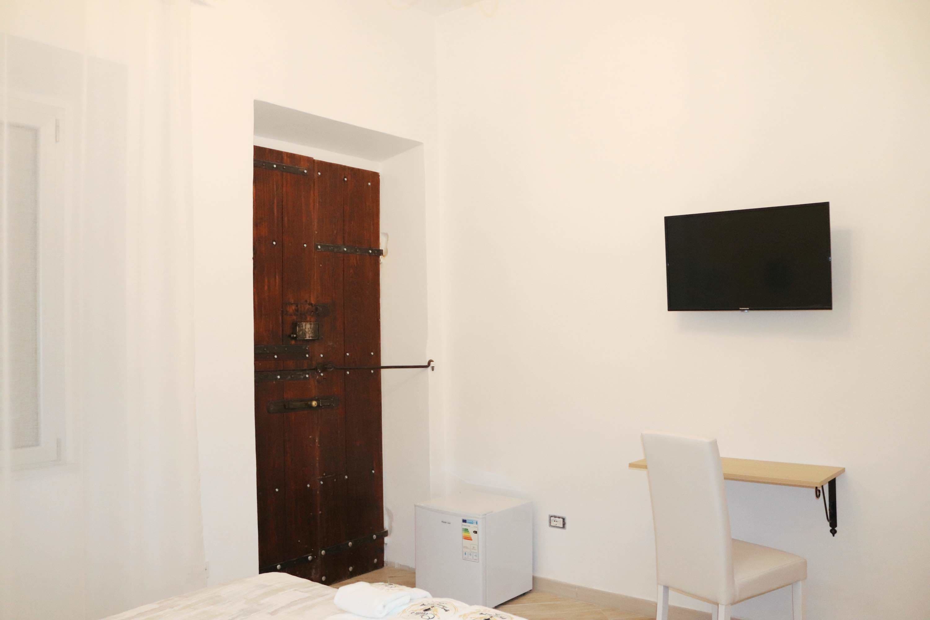 A small room with a wooden door, a white wall-mounted TV, a small white desk, and a bed.