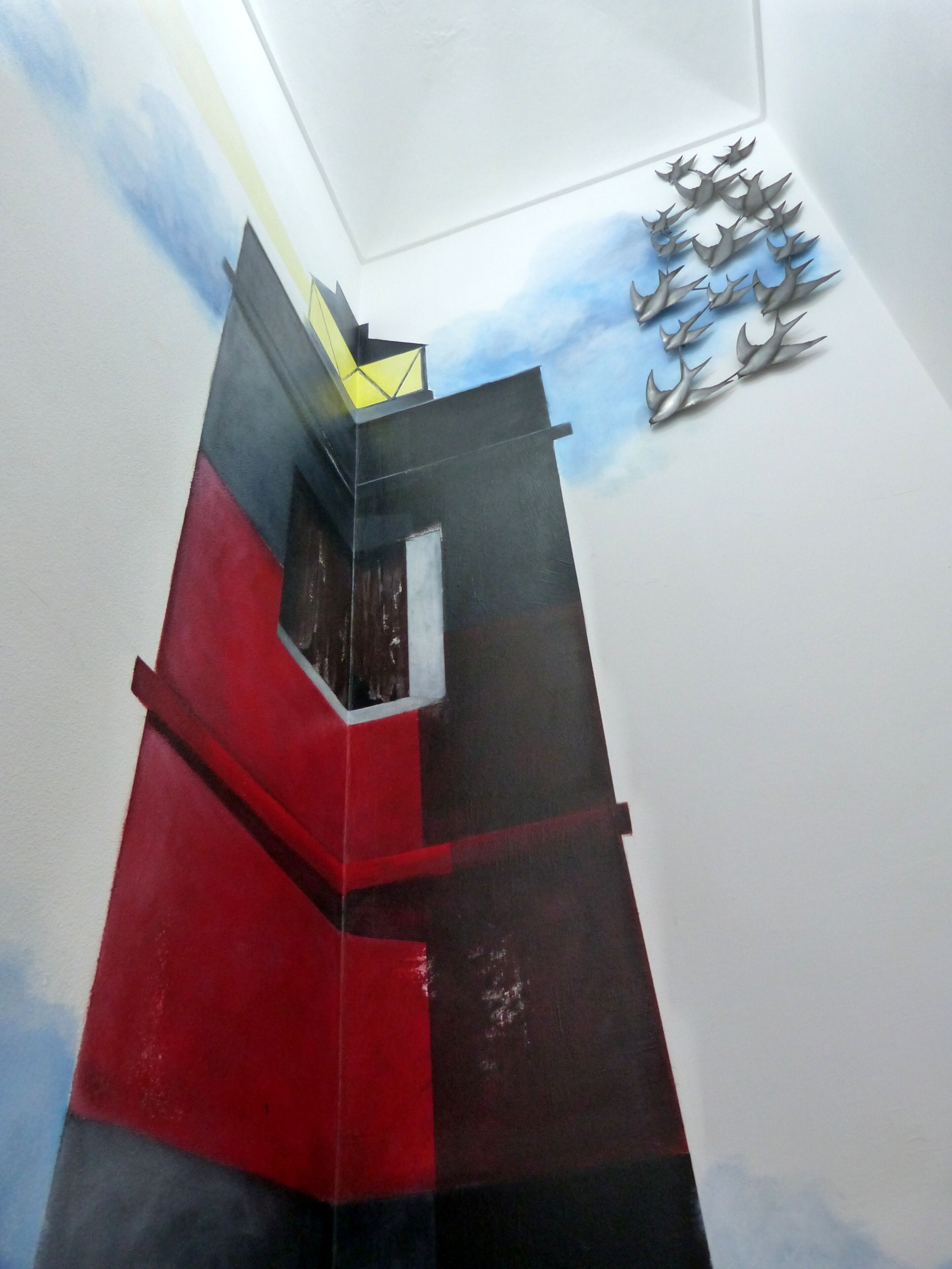 Un edificio moderno con fachada roja y negra, un detalle amarillo en la parte superior y una escultura metálica decorativa a la derecha.
