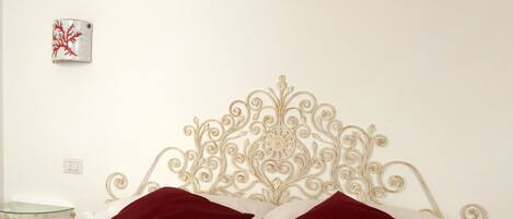 Una cama bien hecha con dos almohadas rojas, un adorno en la pared encima y una mesita de noche con superficie de vidrio al lado.