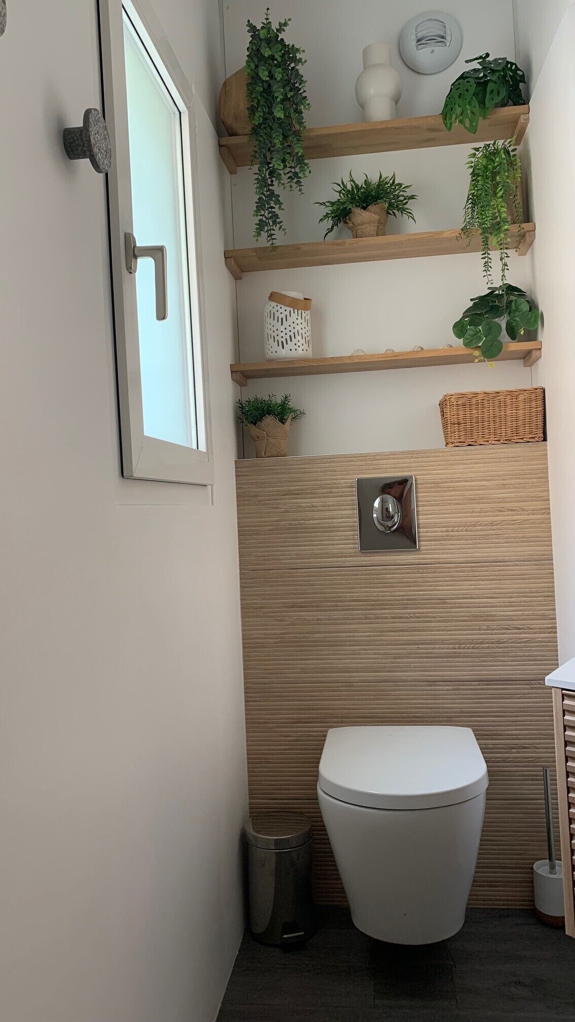 A compact bathroom with a white toilet, a wall-mounted flush handle, and wooden shelves holding plants and decorative items.