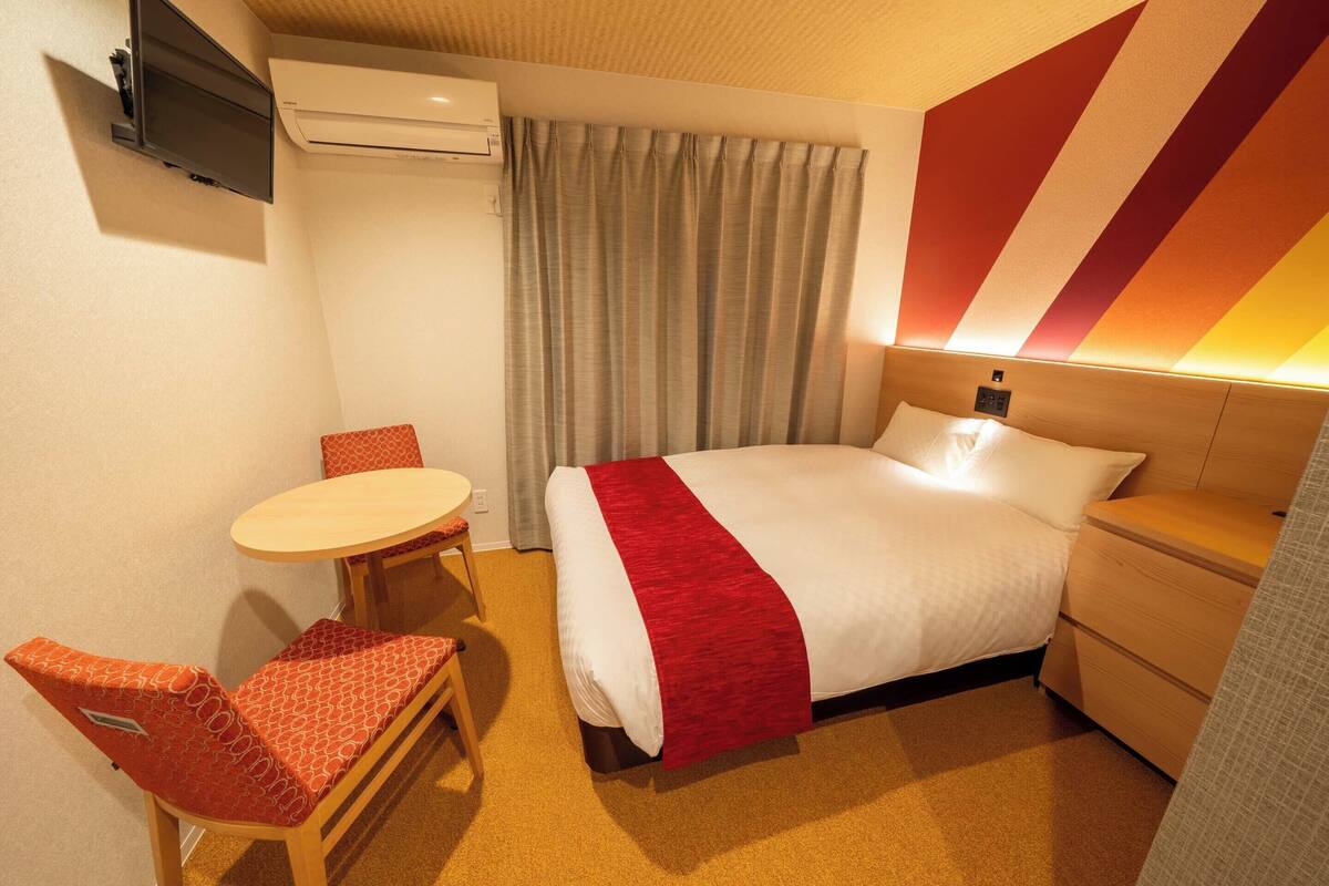 Ein Hotelzimmer mit einem Bett, einem kleinen Tisch mit Stuhl, einer Klimaanlage und einem an der Wand befestigten Fernseher.