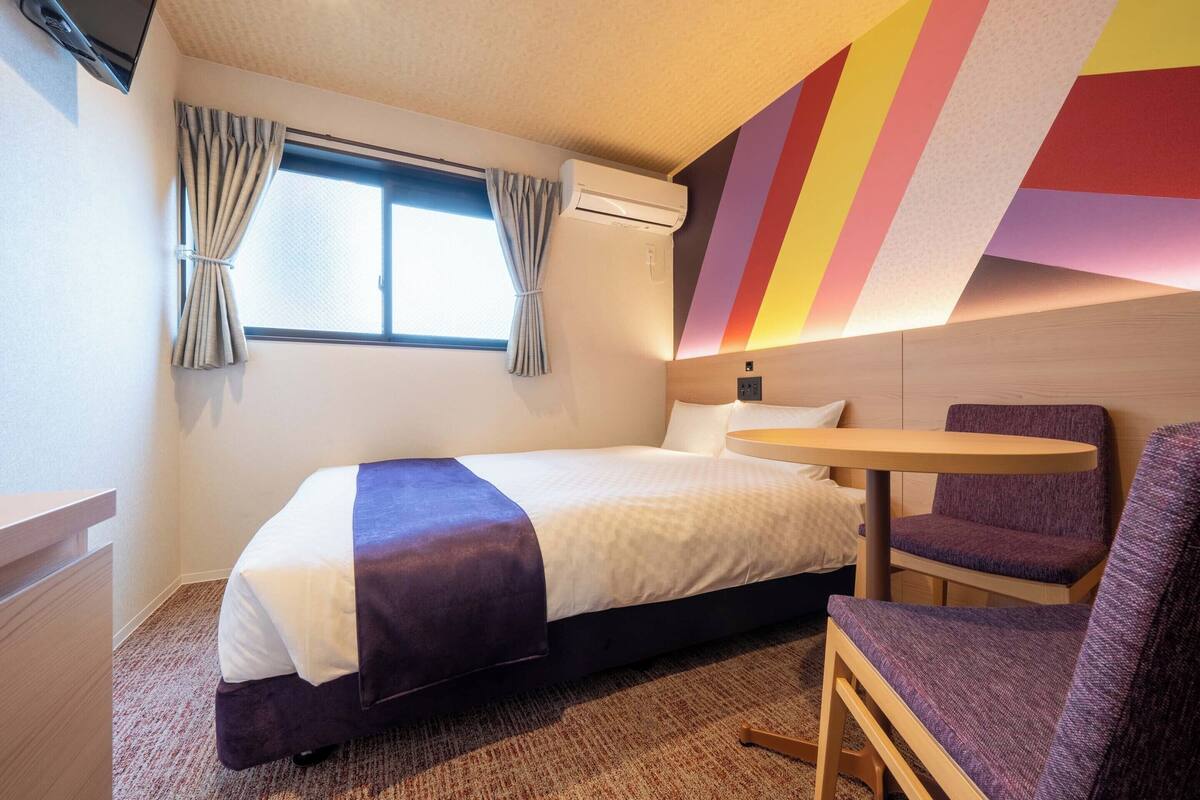 Ein Hotelzimmer mit einem Bett, einem kleinen Tisch, einem Stuhl und einem Fenster mit Vorhängen.
