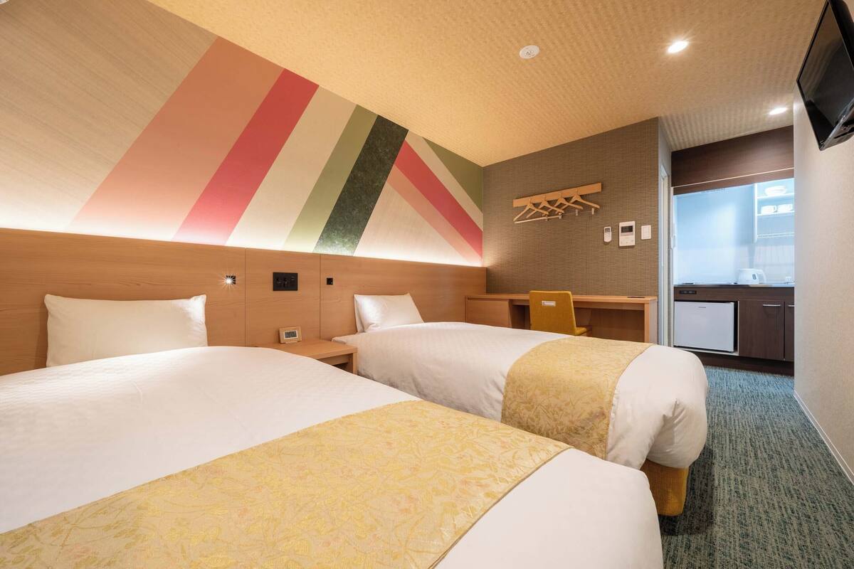 Ein Hotelzimmer mit zwei Betten, einem Schreibtisch, einem Stuhl, einem Fernseher und einer Küchenzeile.