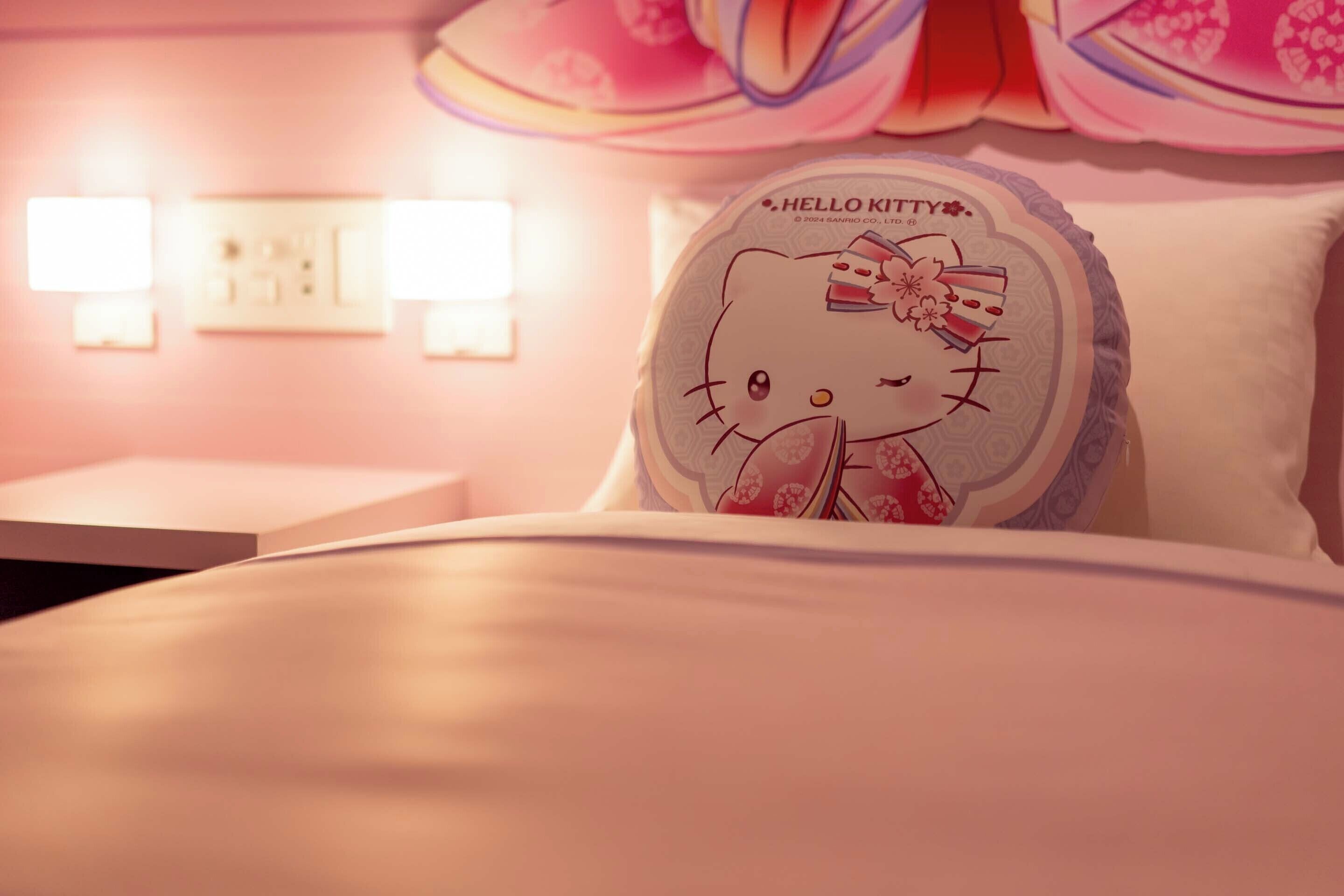 A Hello Kitty-themed pillow on a bed in a hotel room with a pink color scheme and decorative wall sconces.