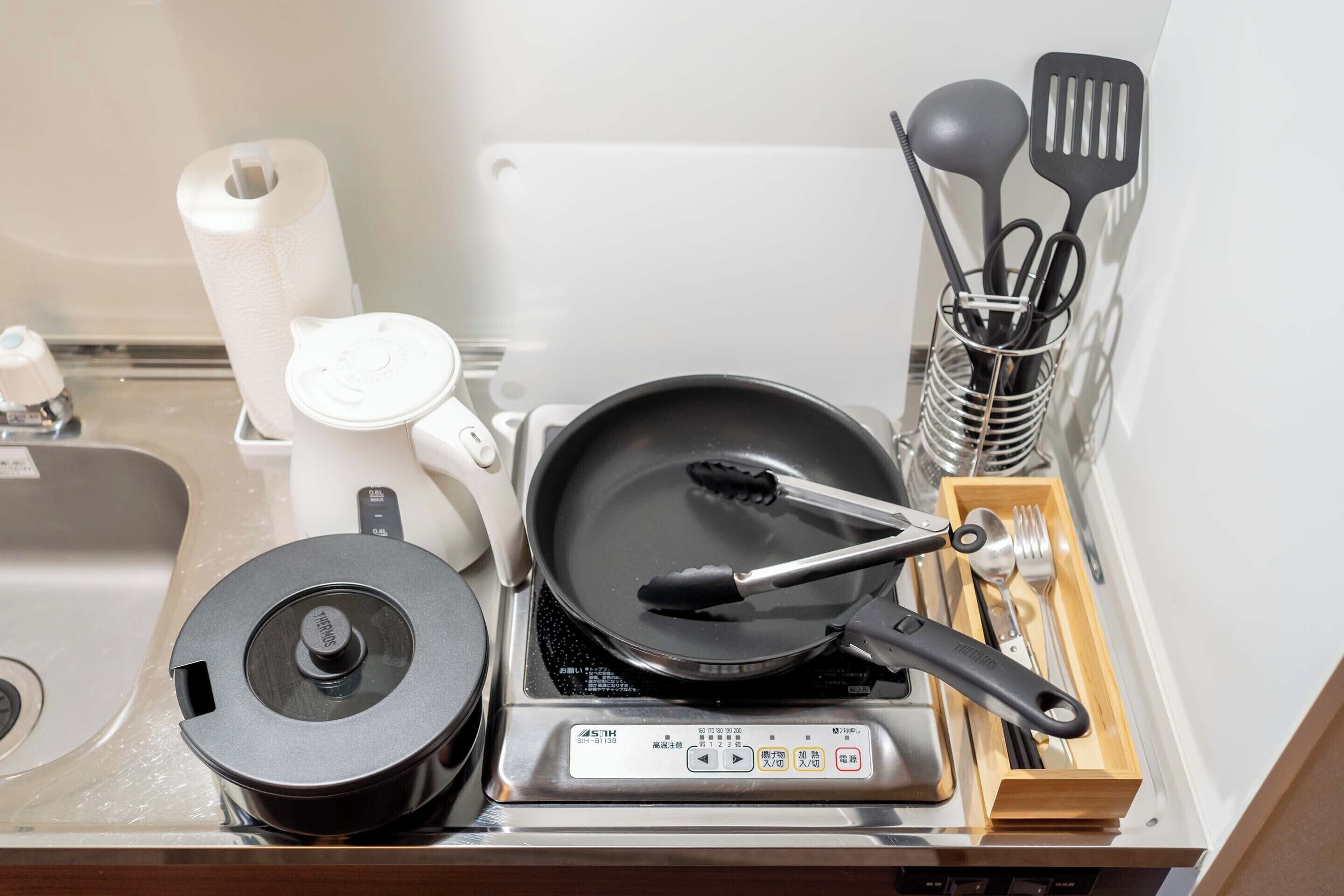 A kitchen area with a stove, a frying pan, a pot, a kettle, and various utensils.