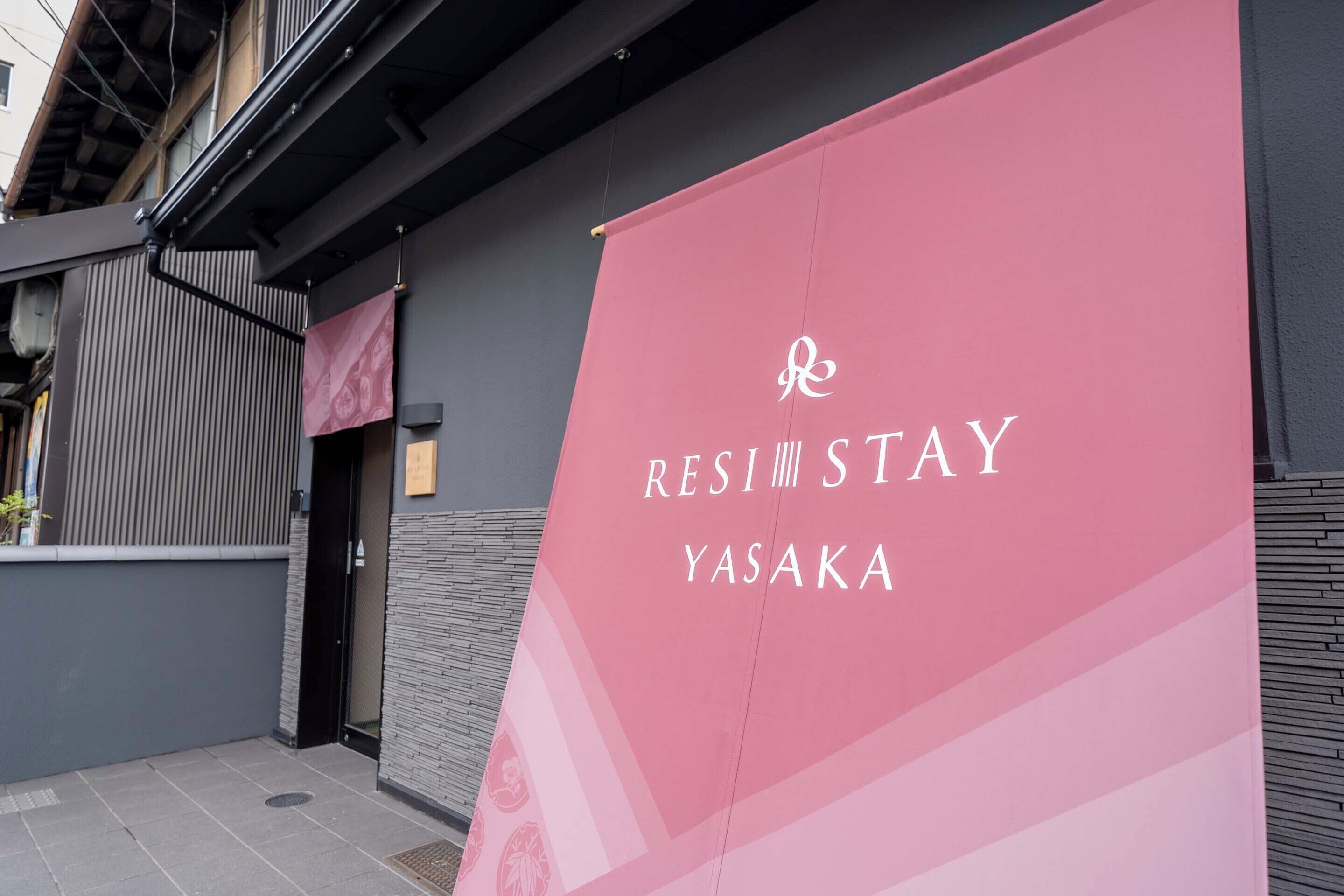 Un banner rosa con el texto "RESI STAY YASAKA", una puerta negra y la fachada de un edificio gris.