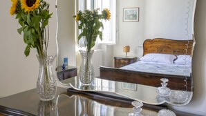 Een slaapkamer met een grote spiegel, een houten hoofdbord en een bed met wit beddengoed. Op een dressoir staan twee glazen vazen met bloemen.