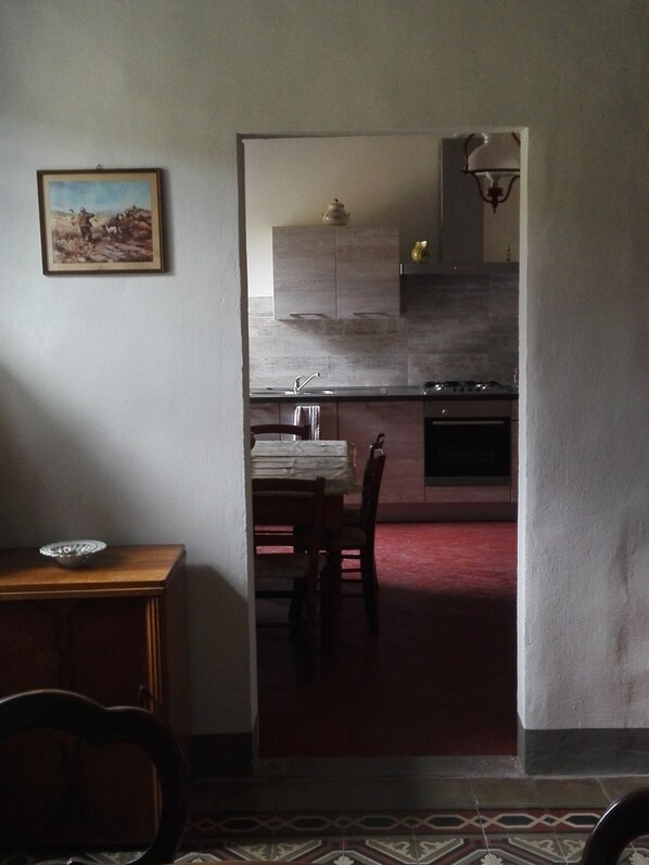 A dining area with a table and chairs, a kitchen in the background with cabinets and a stove, and a painting on the wall. - L'OZioRinco - Floor 1 (Firenze)