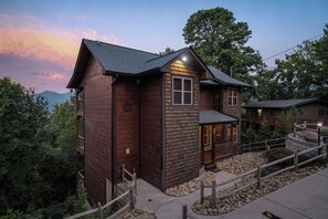 A two-story wooden cabin with a balcony, a stone staircase, and a view of the surrounding forest.