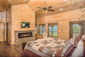 Une chambre dans une cabane en rondins, avec une cheminée en pierre, un lit recouvert d’une couette à carreaux et un ventilateur de plafond.
