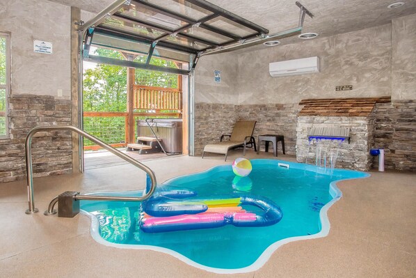 Une piscine intérieure avec un mur en pierre, une terrasse en bois et une porte vitrée donnant sur l’extérieur.