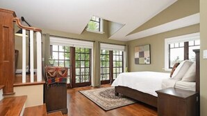 A bedroom with a large bed, wooden flooring, and a view of greenery through the windows. - Character Stay across Swan Lake Nature Sanctuary (Victoria)