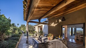 Un área de patio exterior con una pérgola de madera, un bar integrado con una cesta de frutas y vistas del paisaje circundante.