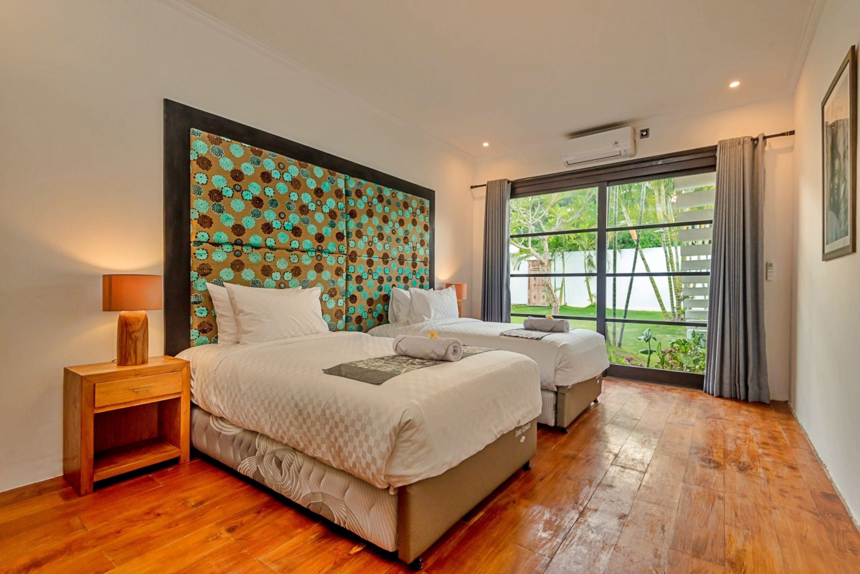 A hotel room with a large bed, a smaller bed, a nightstand, and a window with a view of greenery.