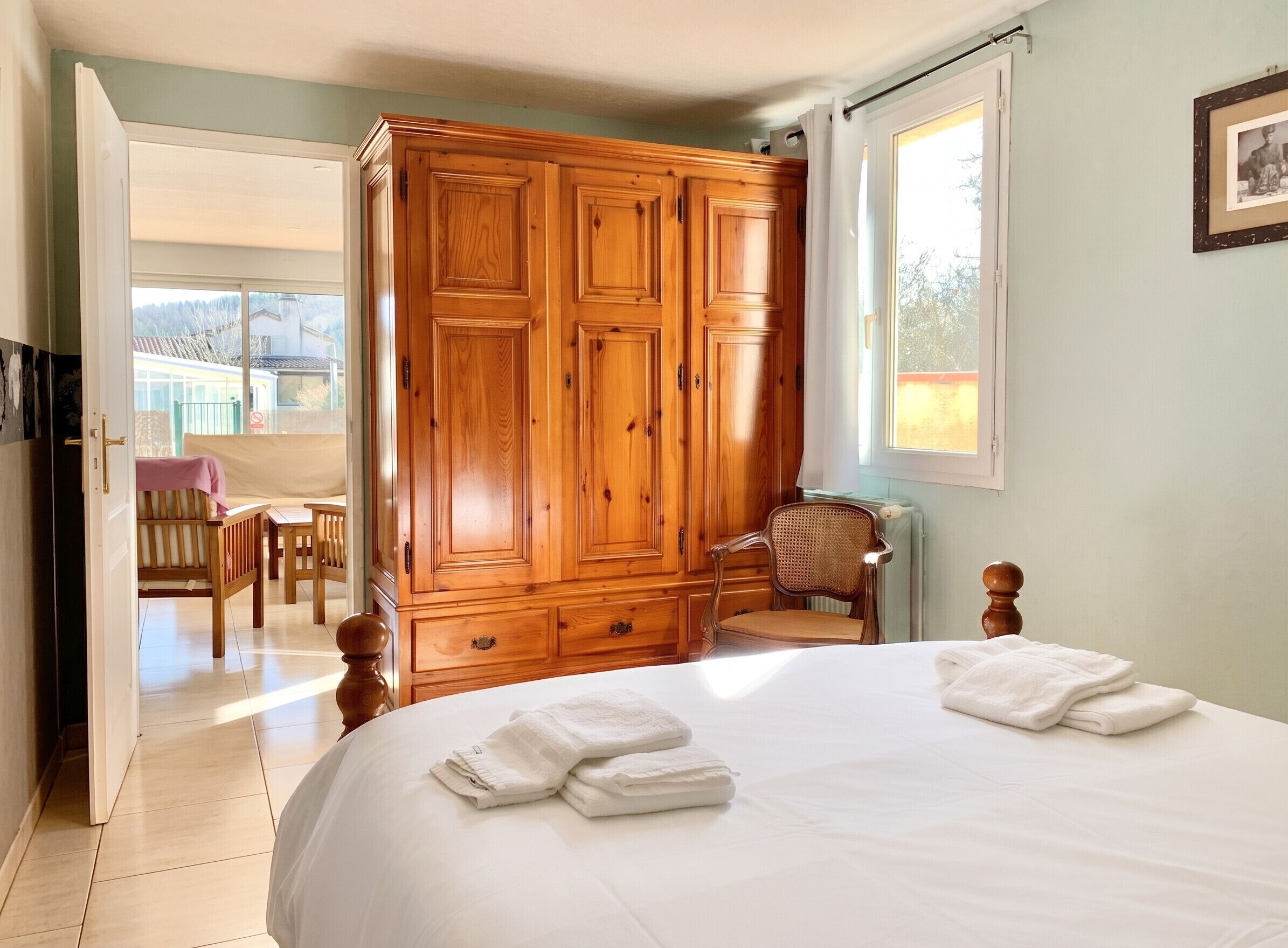 A bedroom with a large wooden wardrobe, a bed with white linens, a chair, and a window.