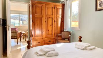 A bedroom with a large wooden wardrobe, a bed with white linens, a chair, and a window.