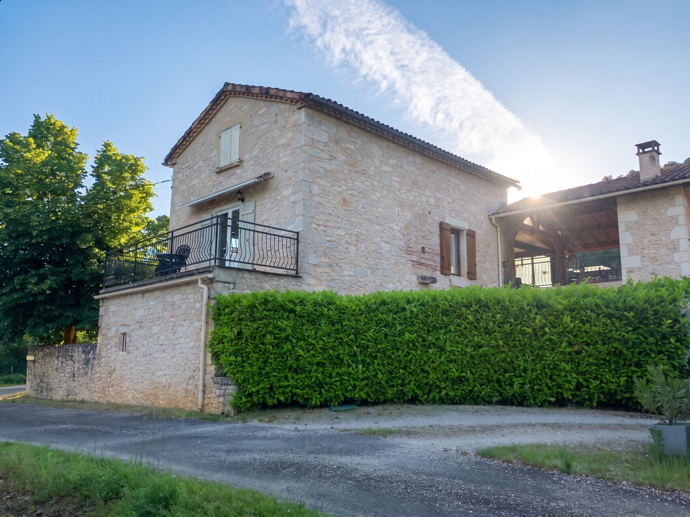 A stone house with a balcony, a hedge in front, and a gravel driveway.