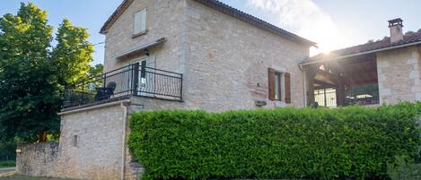 A stone house with a balcony, a hedge in front, and a gravel driveway.