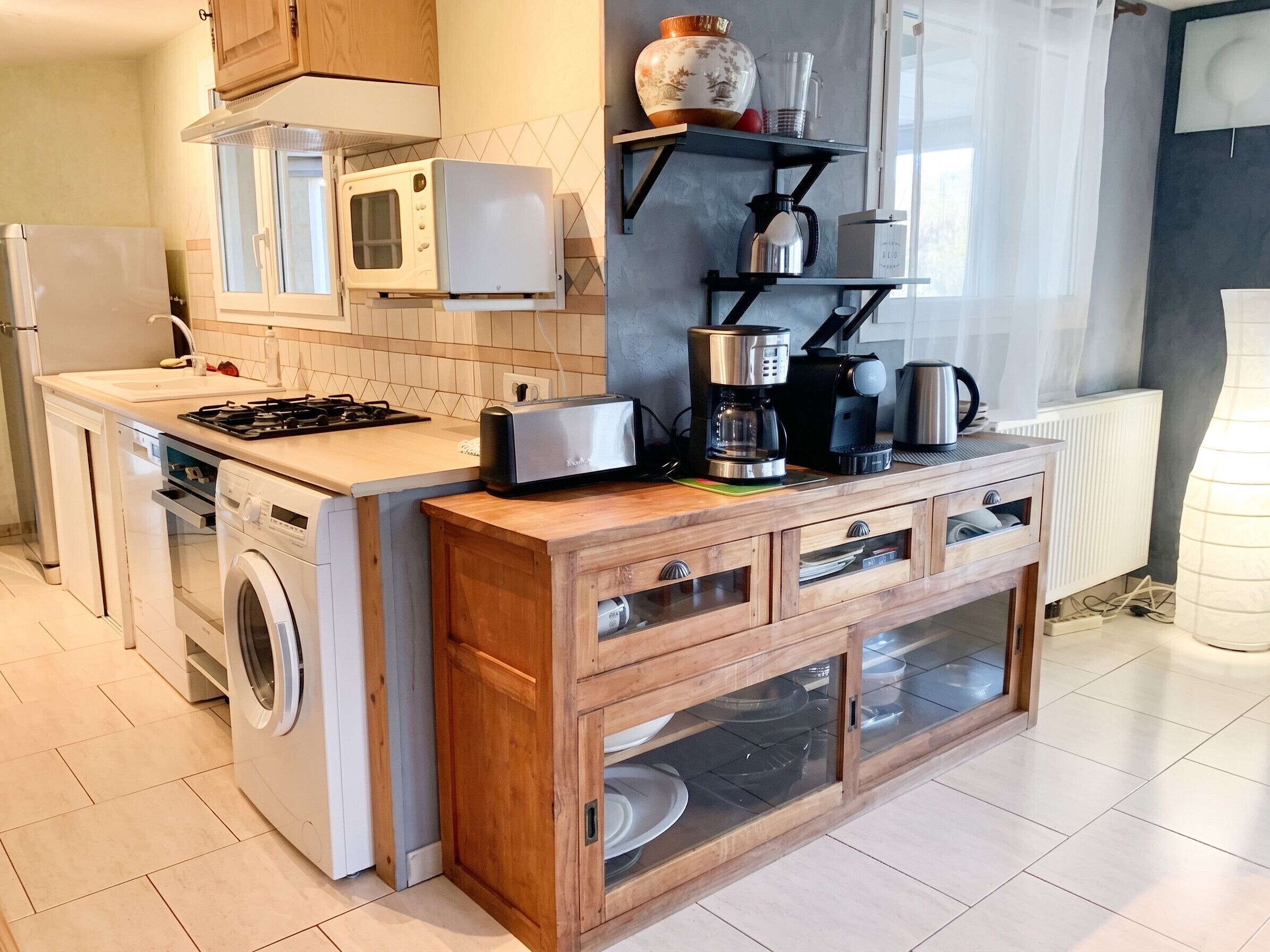 A compact kitchen with a washing machine, microwave, toaster, coffee maker, and a cabinet with glass doors.