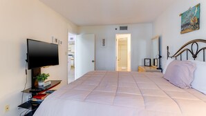 Um quarto com uma cama grande, uma TV em um suporte, uma mesa de cabeceira com abajur e uma porta que dá para outro cômodo.