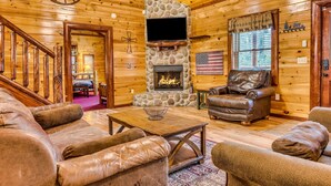 A log cabin interior with a stone fireplace, leather furniture, and a wooden staircase. - Heated Pool, Movie Room, Sleeps 30, Dogs Welcome (Cosby)