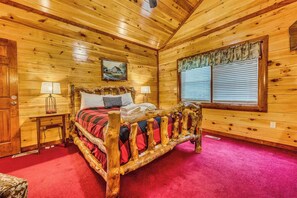 A wooden cabin bedroom with a bed, two bedside tables, a window with blinds, and a painting on the wall. - Pools Rush Inn (Cosby)