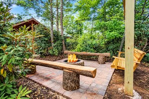 An outdoor fire pit area with a swing chair, surrounded by greenery and a brick pathway. - Mountain Retreat • Sleeps29 • Pool • HotTub • Dogs (Cosby)