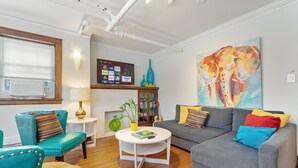 Ein Wohnzimmer mit türkisfarbenen Sesseln, einem grauen Sofa, einem runden Couchtisch und einem farbenfrohen Elefantenbild an der Wand.