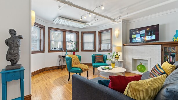 Ein modernes Wohnzimmer mit Kamin, smaragdgrünen Sesseln, einem grauen Sofa und einem Flachbildfernseher, der Streaming-Dienste anzeigt.