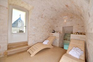 A stone-walled room with two wooden loungers, a small shelf with items, and a view of a building outside.