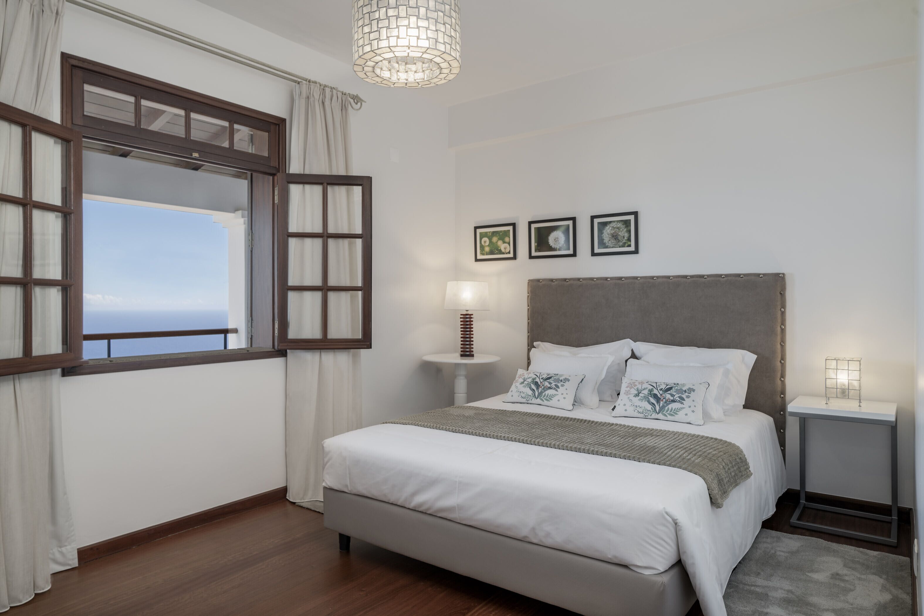A bedroom with a large bed, a bedside table, a lamp, and a view of the sea through a window with curtains.