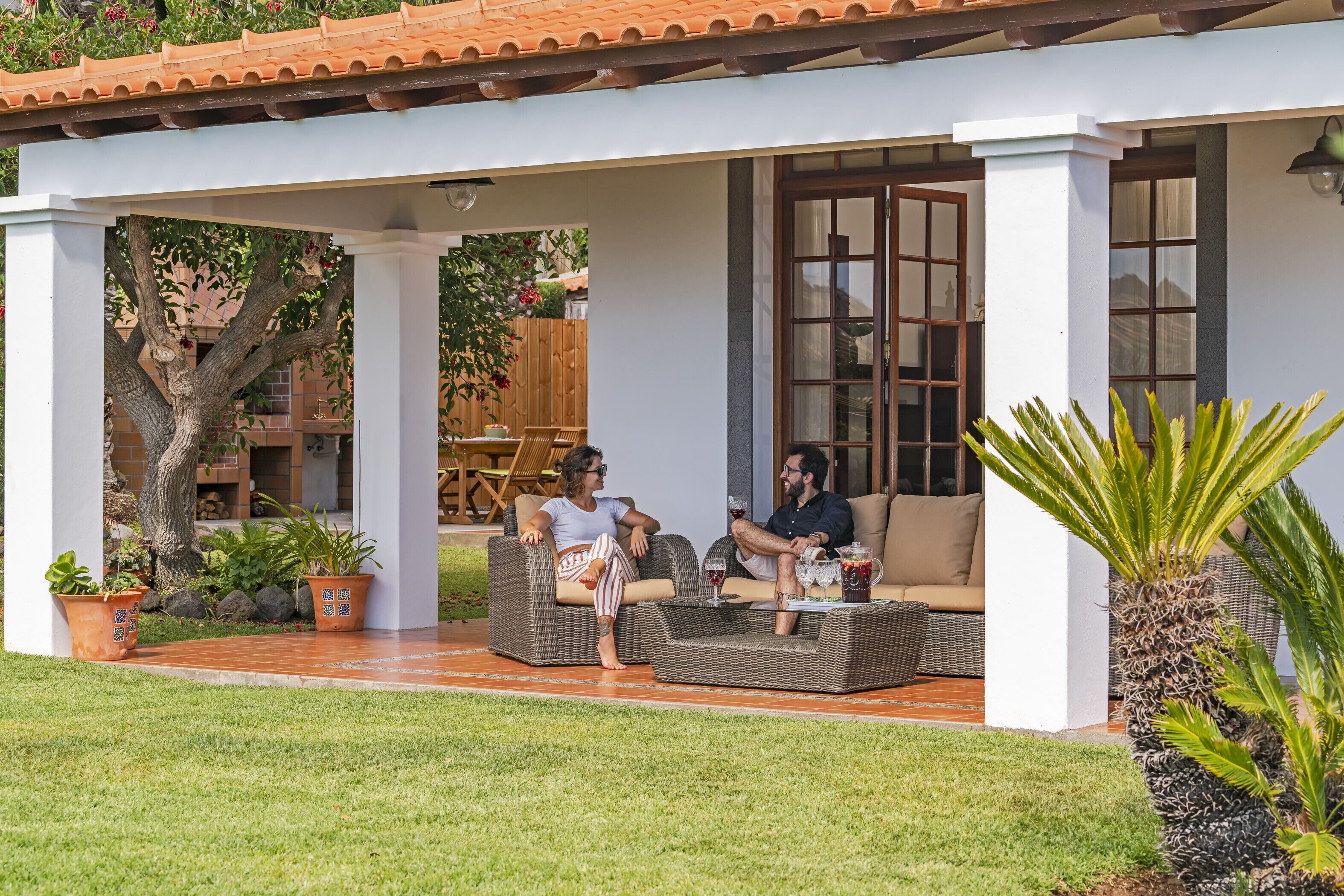 A covered outdoor seating area with wicker furniture, potted plants, and a view of a building in the background.